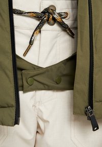 Gros plan d'un pantalon beige avec une ceinture en corde multicolore et une veste vert olive dézippée montrant des boutons-pression en plastique et une tirette de fermeture éclair.