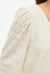 Kremowa bluzka z teksturowanej tkaniny z diamentowym wzorem. Bufiaste rękawy dodają objętości, a dekolt w kształcie litery V.
