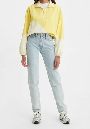Personne portant un jean taille haute bleu clair, des baskets blanches et un pull ample jaune et crème à manches longues avec une fermeture éclair sur le devant.