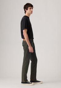 Joven de pie de lado, vistiendo una camiseta negra, jeans verde oscuro y zapatillas negras, contra un fondo claro y uniforme.