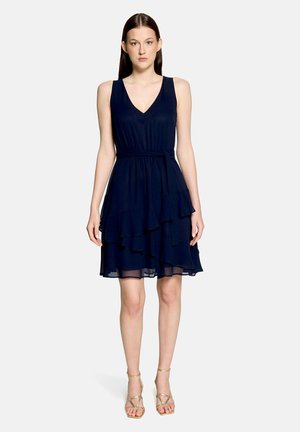 Kobieta nosząca bezrękawową granatową sukienkę z dekoltem w serek, z warstwową spódnicą i przewiązanym paskiem w talii, w połączeniu ze złotymi sandałami na paski, stojąca prosto.