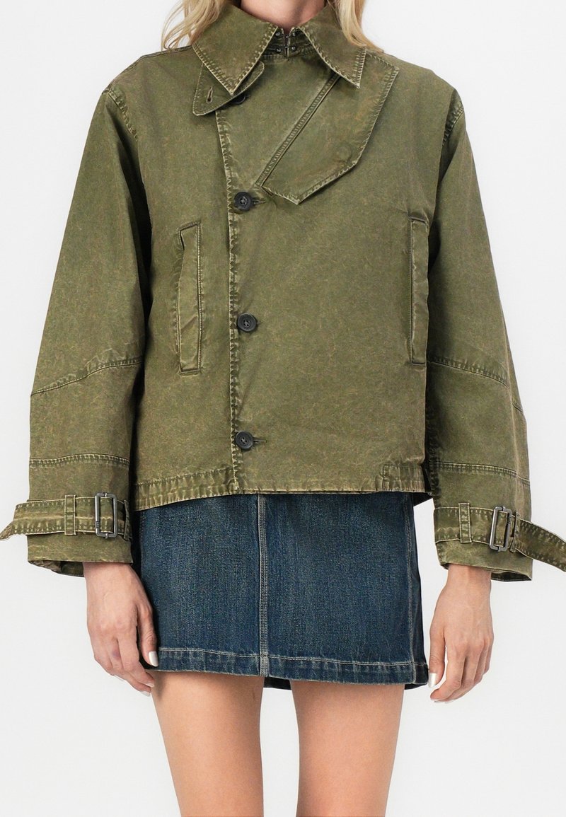 Veste courte vert olive à fermeture asymétrique par boutons, sangles à boucle aux poignets, associée à une mini-jupe en jean bleue.