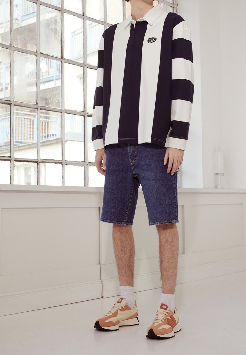 Polo à manches longues avec larges rayures bleu marine et blanches, assorti à un short en jean mi-long et des baskets multicolores.