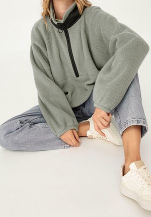 Femme assise portant un pull polaire vert clair avec une fermeture éclair noire, un jean gris clair et des baskets blanches sur un fond uni.