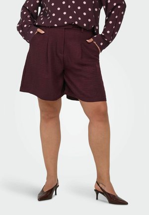 Žena nosiaca tmavé bordové krátke nohavice na mieru s rukami v vreckách, v kombinácii s blúzkou s bodkami a tmavohnedými podpätkami.