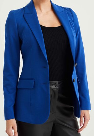 Femme portant un blazer bleu ajusté sur un haut côtelé noir et un pantalon en cuir noir, debout devant un fond clair uni.