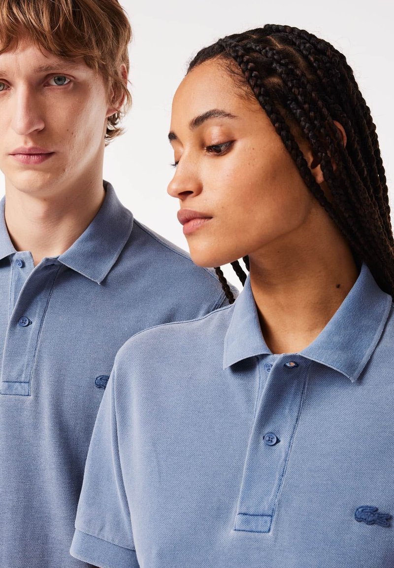 Dos personas que llevan camisetas polo de color azul claro, hechas de un tejido texturizado. Las camisetas tienen un cuello y un pequeño logotipo bordado en el pecho.