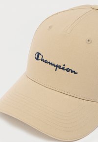 Μπεζ καπέλο μπέιζμπολ κατασκευασμένο από υφασμάτινη υφή με κυρτή βρεχόμενη και κεντημένο μπλε ναυτικό λογότυπο "Champion" στο μπροστινό μέρος.