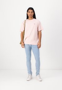 Camiseta de algodón de color rosa claro con mangas cortas y un pequeño logo en el pecho, combinada con jeans ajustados de color azul claro y zapatillas blancas.