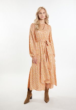 Langes, gemustertes Kleid in Orange und Creme mit einem Taillengürtel und langen Ärmeln. Dazu werden braune Stiefeletten getragen.