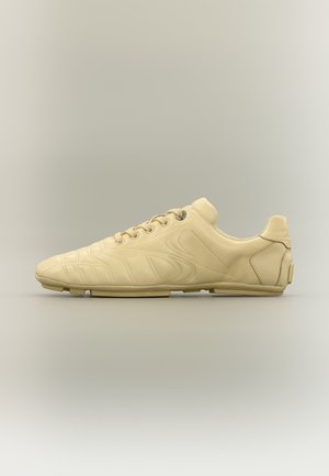 Beiger minimalistischer Sneaker mit Schnürverschluss, dezenten geprägten Mustern an der Seite und flacher Sohle, von der Außenseite auf neutralem Hintergrund gezeigt.