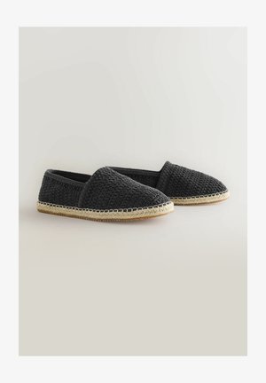 Melnas austas slip-on kurpes ar faktūru augšdaļu, ar jutu zoli un šuvju detaļām malā. Plakans, ikdienišķs dizains.