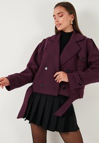 Purpurna dvoredna jakna iz teksturiranega materiala, ki ima velike gumbe, širok ovratnik in pas z obročem, v kombinaciji s črno plisirano krilo.