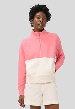 Vrouw met kort krullend haar, gekleed in een roze-witte trui met een kwart rits en witte korte broek, staand met één hand in de zak.