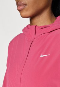 Veste Nike rose avec fermeture éclair sur le devant, capuche et logo réfléchissant. Texture lisse avec un minimum de détails de couture et une conception ajustée.