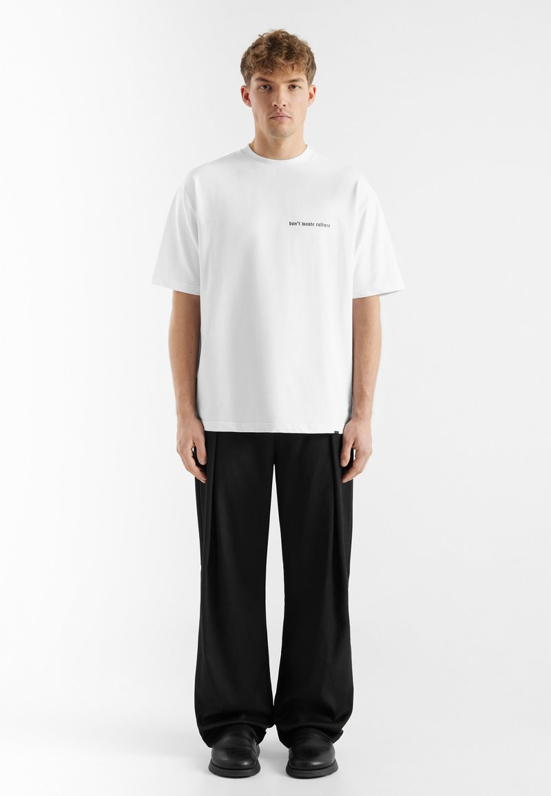 Witte katoenen t-shirt met korte mouwen, met zwarte tekst "verspilt cultuur" op de voorkant. Gecombineerd met losse zwarte broek en zwarte schoenen.