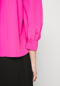 Fuksia pusero, jossa on rento malli, kerätty hihna ja napillinen hihansuu. Pusero on valmistettu sileästä, teksturoidusta kankaasta, ja siinä on kaareva helma.