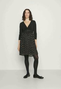 Fekete ruha hosszú ujjaival és mély dekoltázzsal, pöttyös mintával átlátszó szoknyán. Fekete harisnyával és lapos cipővel viselve.