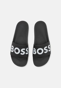 Fekete csúszó szandál texturált talpbetéttel és széles fekete pánttal, melyen vastag, fehér betűkkel szerepel a "BOSS" felirat. Lapos, nyitott orrú kialakítás.