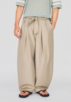 Homme portant un pantalon beige à jambes larges avec une ceinture nouée, un haut en tricot vert clair et des sandales noires, debout les mains dans les poches.