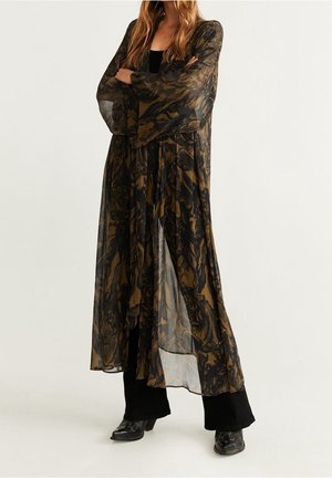 Femme portant un long cardigan transparent noir et marron à motif style kimono, sur un pantalon noir et des chaussures noires, debout bras croisés.