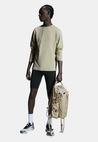 Beige overhemd met lange mouwen gecombineerd met zwarte shorts. De persoon houdt een tan rugzak vast met meerdere vakken en riemen. Zwarte en grijze sneakers.
