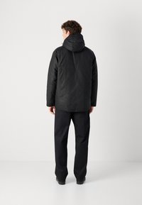 Fekete kapucnis dzseki matt felülettel, laza szabással, hosszú ujjakkal és strukturált anyagból készült, sötét nadrággal és cipővel párosítva.