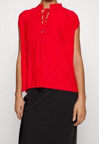 Rød plisseret top med høj hals, knyttet nøglehul detalje og korte ærmer, parret med en tætsiddende sort nederdel. Glat stofstruktur.