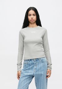 Νεαρή γυναίκα με μακριά μαύρα μαλλιά, φορώντας γκρι κορυφή με ριμπ και μακριά μανίκια με το λογότυπο "Jordan" και ανοιχτό μπλε ριγέ τζιν.