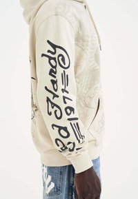 Sudadera beige con texto gráfico negro en las mangas, con un diseño texturizado y un bolsillo frontal. Los puños y el dobladillo de canalé completan el look.