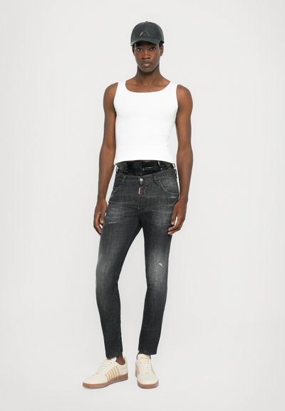 Jeans de mezclilla negros para hombre con textura desgastada, combinados con una camiseta sin mangas blanca ajustada y zapatillas crema. Una gorra negra completa el atuendo.