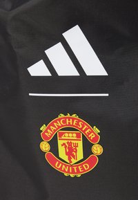 A fekete anyagon a bal felső sarokban egy hangsúlyos fehér Adidas logó, az alsó jobb sarokban pedig egy piros és sárga Manchester United címer található.