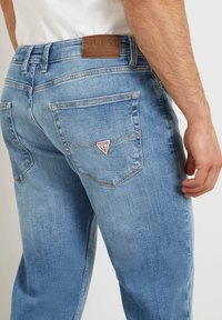 Vaqueros de mezclilla azul claro que presentan un bolsillo con logo triangular bordado, un parche de cuero en la cintura y un diseño clásico de cinco bolsillos.