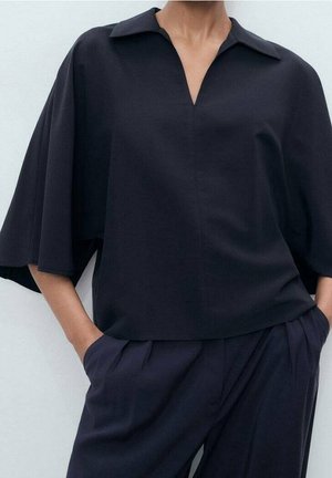 Femme portant un haut noir ample à col en V avec des manches larges et un pantalon noir plissé assorti, mains dans les poches, sur un fond uni.