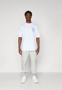 Hvit T-skjorte med korte ermer og blått grafisk design på brystet, kombinert med lysegrå cargobukser og hvite treningssko.