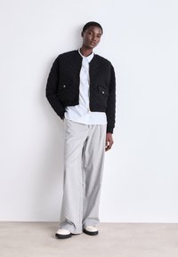 Casaco bomber acolchoado preto com detalhes em dourado, combinado com uma camisa de botão azul clara e calças largas cinza claro. Sapatos brancos e pretos.