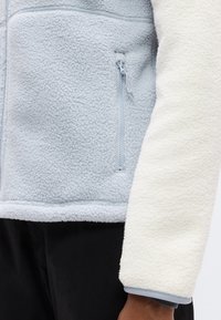 Ανοιχτό μπλε fleece μπουφάν με λευκό fleece μανίκι, που διαθέτει τσέπη με φερμουάρ, υφή υφάσματος και απαλό, άνετο στυλ.