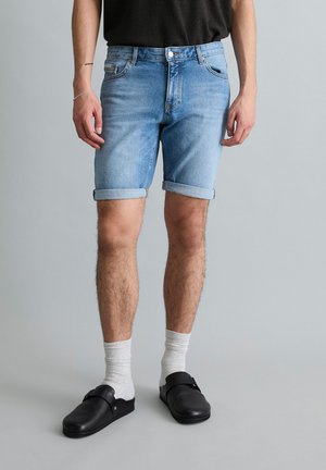 Man wearing rolled-up light blue denim shorts, white socks, black slip-on shoes, and a black shirt, standing against plain gray background.