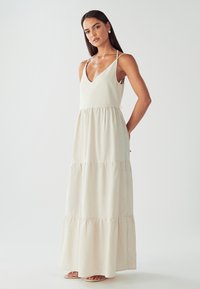 Moteris, vilkinti be rankovių, tiered smėlio spalvos maxi suknelę su plonomis petnešomis ir V formos iškirpte, stovi su rankomis už nugaros.