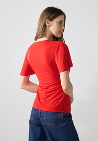 Top scurt roșu cu mâneci scurte și guler rotund, având un design ajustat și detalii pliate la spate; asortat cu blugi din denim blue cu picior larg.