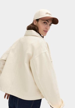 Vrouw met een beige oversized jas en een tweekleurige honkbalpet, kijkt over haar schouder terug tegen een effen achtergrond.