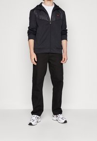 Black zip-up hoodie with textured dark upper panel, worn over a white t-shirt, paired with black pants and white sneakers.