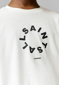 Bela bombažna majica s velikim črnim besedilom v krožnem dizajnu, z "SAINT" na vrhu, "LONDON" centrirano spodaj in krepko napisanimi črkami.
