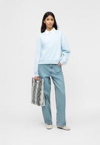 Γυναίκα που φοράει ανοιχτό μπλε φούτερ πάνω από λευκό πουκάμισο με γιακά, τζιν παντελόνι με φαρδιές φάσες, μπεζ παπούτσια, κρατώντας μια μεγάλη τσάντα με μοτίβο δέρματος φιδιού.