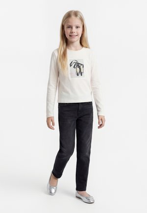 Mladé dievča s dlhými blond vlasmi má na sebe biele tričko s dlhým rukávom s potlačou baletných topánok, čierne rifle a strieborné balerínky.