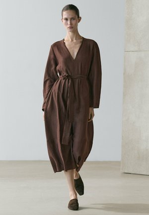 Woman wearing a long-sleeve, belted, dark brown midi dress walking indoors with matching dark brown slip-on shoes.