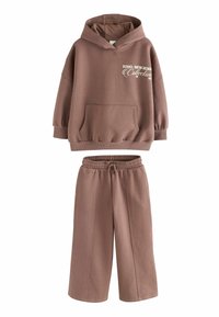 Hoodie in cotone marrone con una grande tasca frontale e design testuale bianco, abbinato a pantaloni larghi coordinati con una vita elasticizzata.