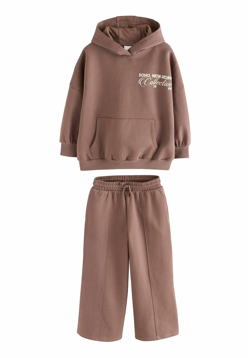 Hoodie in cotone marrone con una grande tasca frontale e design testuale bianco, abbinato a pantaloni larghi coordinati con una vita elasticizzata.