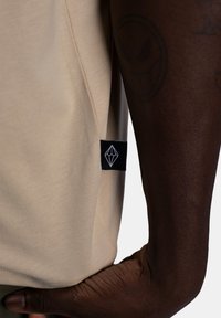 Beżowa bawełniana koszulka z gładką teksturą, z czarną metką z białym geometrycznym logo po lewej stronie szwu.