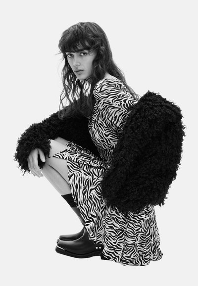 Mujer de cabello largo agachada, vestida con un vestido de estampado de cebra, abrigo negro texturizado sobre los hombros, botas negras hasta el tobillo y calcetines negros.